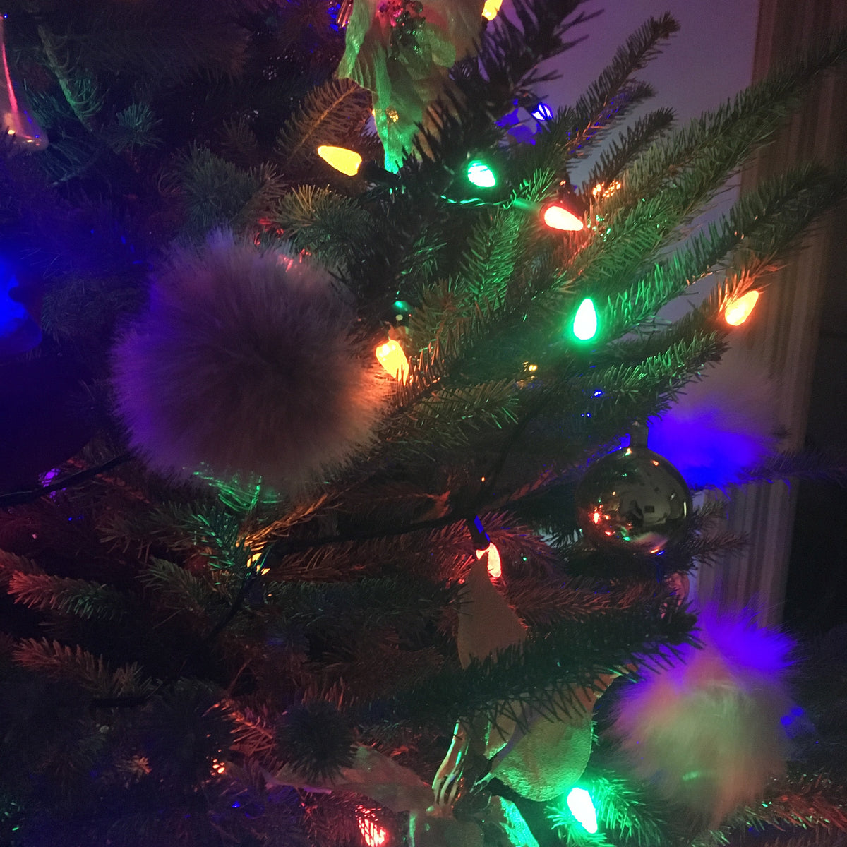 A close-up of a decorated Christmas tree with green artificial needles, multicolored string lights in red, green, yellow, and blue, fluffy white pom-poms, and a small reflective ornament.