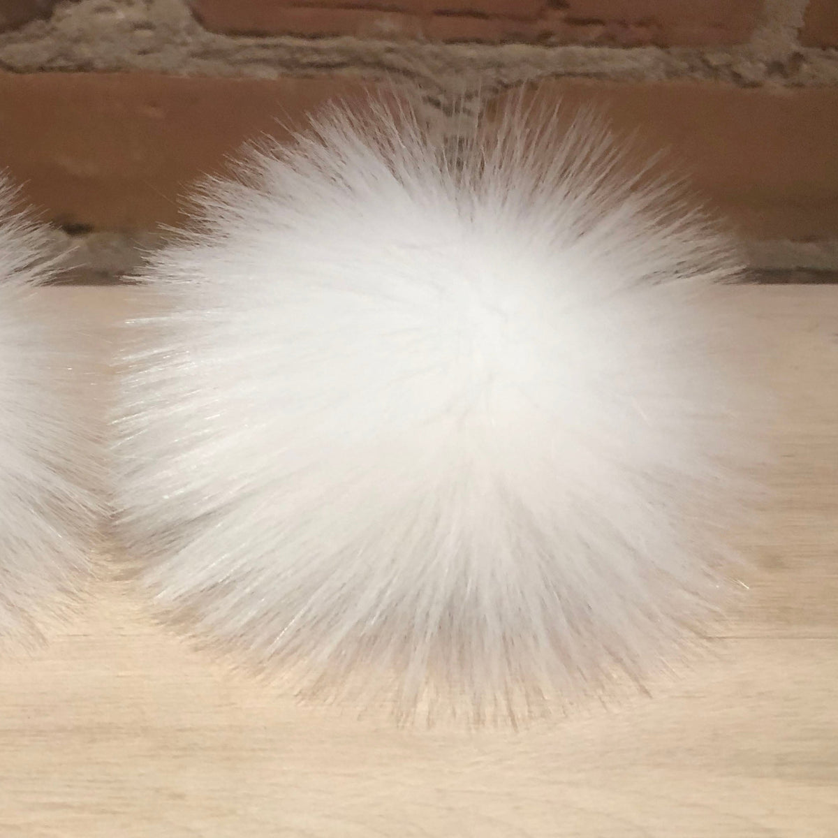 Two fluffy white faux fur pom-poms resting on a light wood surface in front of a brick wall background.