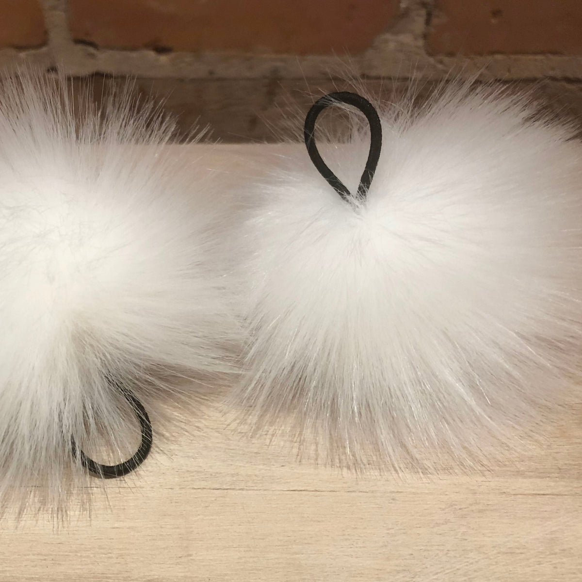 Two fluffy white faux-fur pom-pom accessories with black hanging loops, displayed on a light wood surface against a brick wall background.
