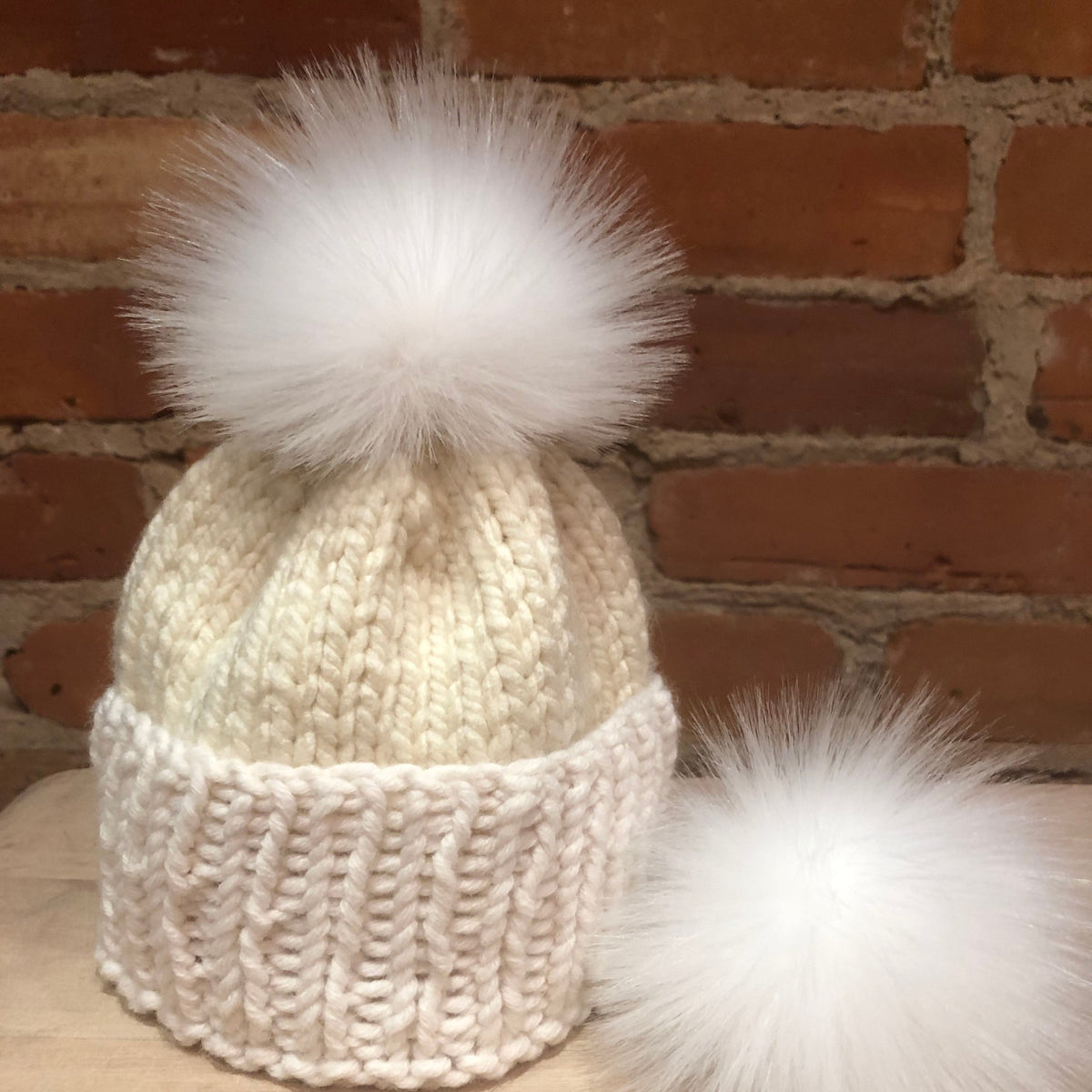 A cream-colored knit beanie hat with a chunky ribbed brim and a large fluffy white faux-fur pom-pom on top, displayed on a wooden surface against a brick wall background.