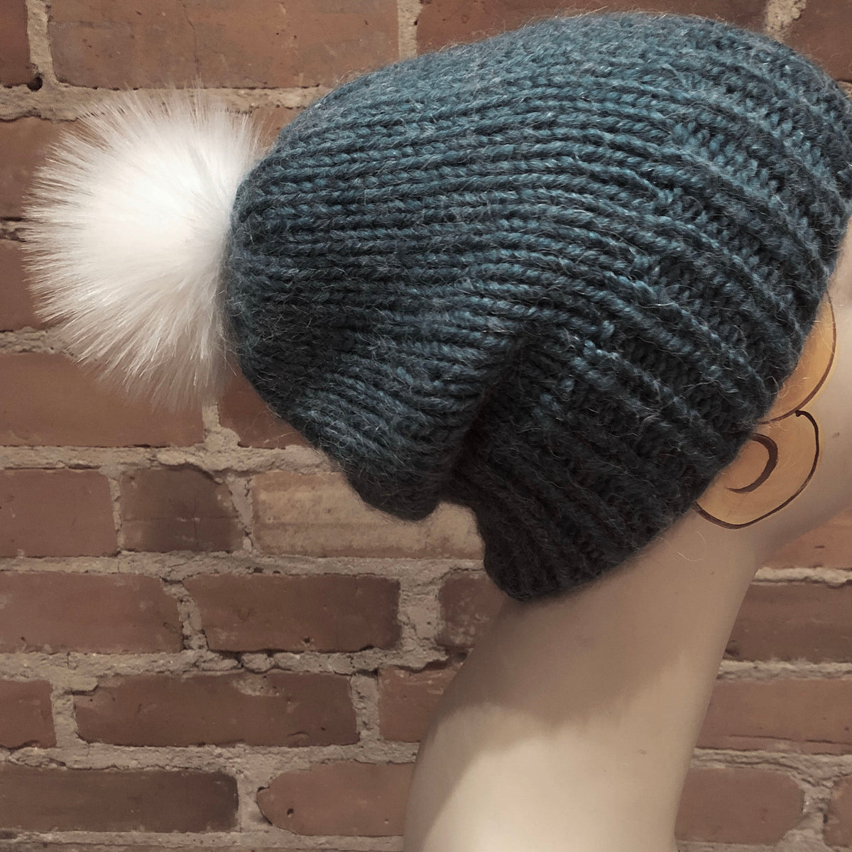 A dark teal blue-green chunky knit slouchy beanie with a fluffy white faux fur pom-pom, displayed on a mannequin head against a brick wall background.
