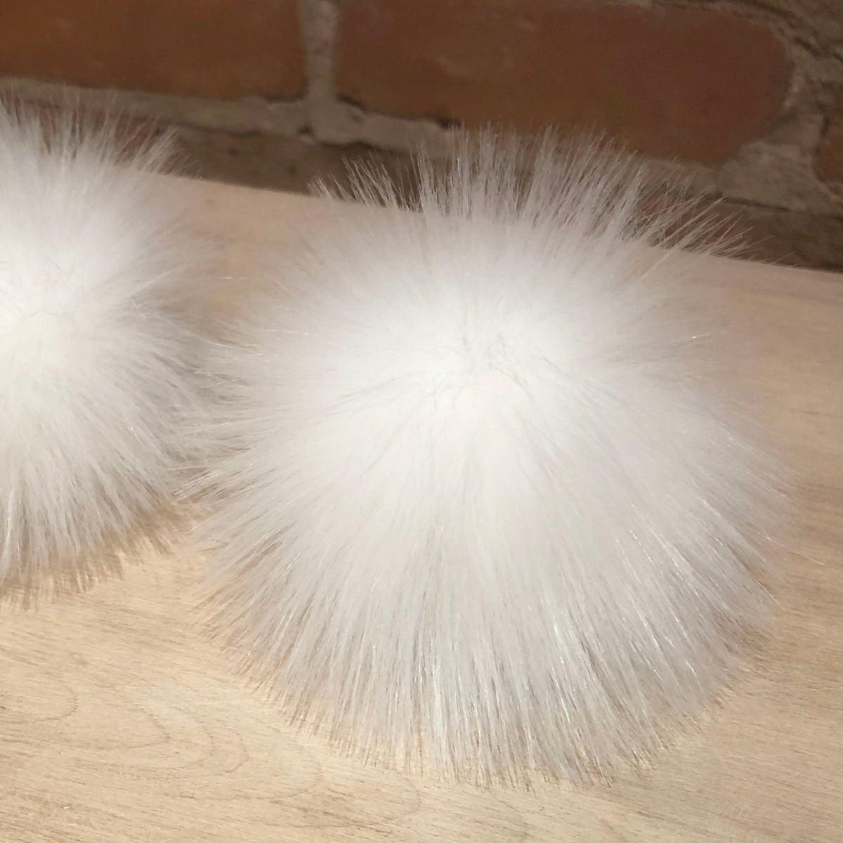 Two fluffy white faux-fur pom-poms resting on a light wood surface with a brick wall in the background.