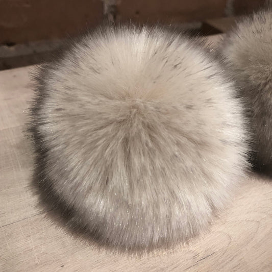 A close-up of a fluffy faux fur pom-pom in a light gray and white colorway, resting on a light wood surface.