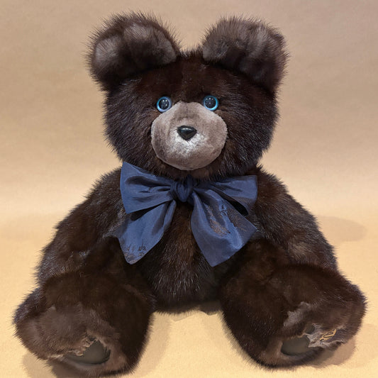 A dark brown plush mink teddy bear with a gray snout, blue eyes, and a dark blue satin bow tie, sitting against a plain beige background.