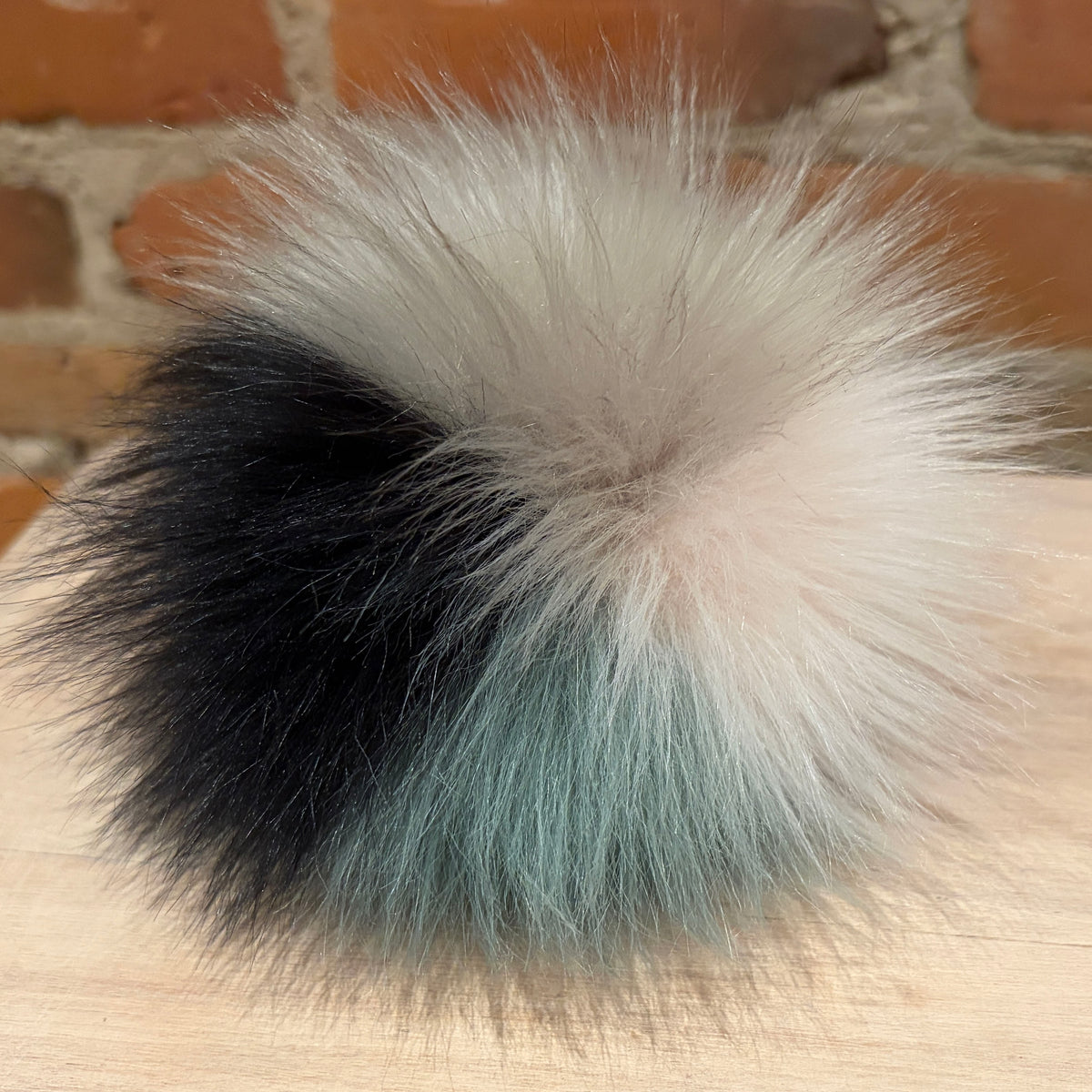 A fluffy faux-fur pom pom accessory with a color-blocked design in black, cream, and light blue, resting on a light wood surface with a brick wall background.