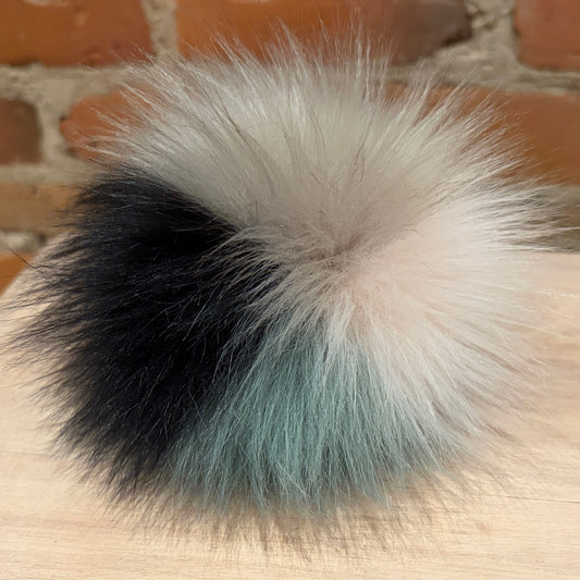 A fluffy faux-fur pom pom accessory with a color-blocked design in black, cream, and light blue, resting on a light wood surface with a brick wall background.