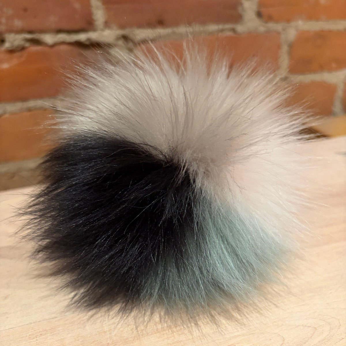 A fluffy pom-pom with a three-color gradient of black, light blue, and cream, resting on a light wood surface with a brick wall background.