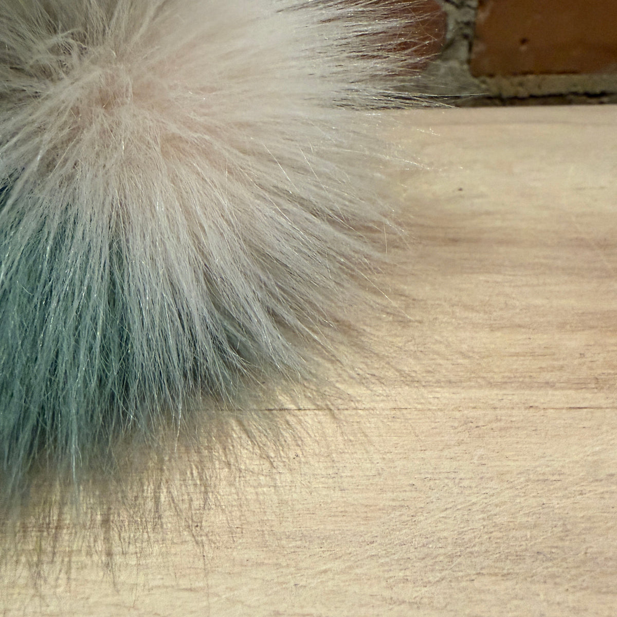 A close-up of a fluffy faux fur pom-pom accessory with a soft cream top fading into pale blue-green at the bottom, resting on a light wood surface.