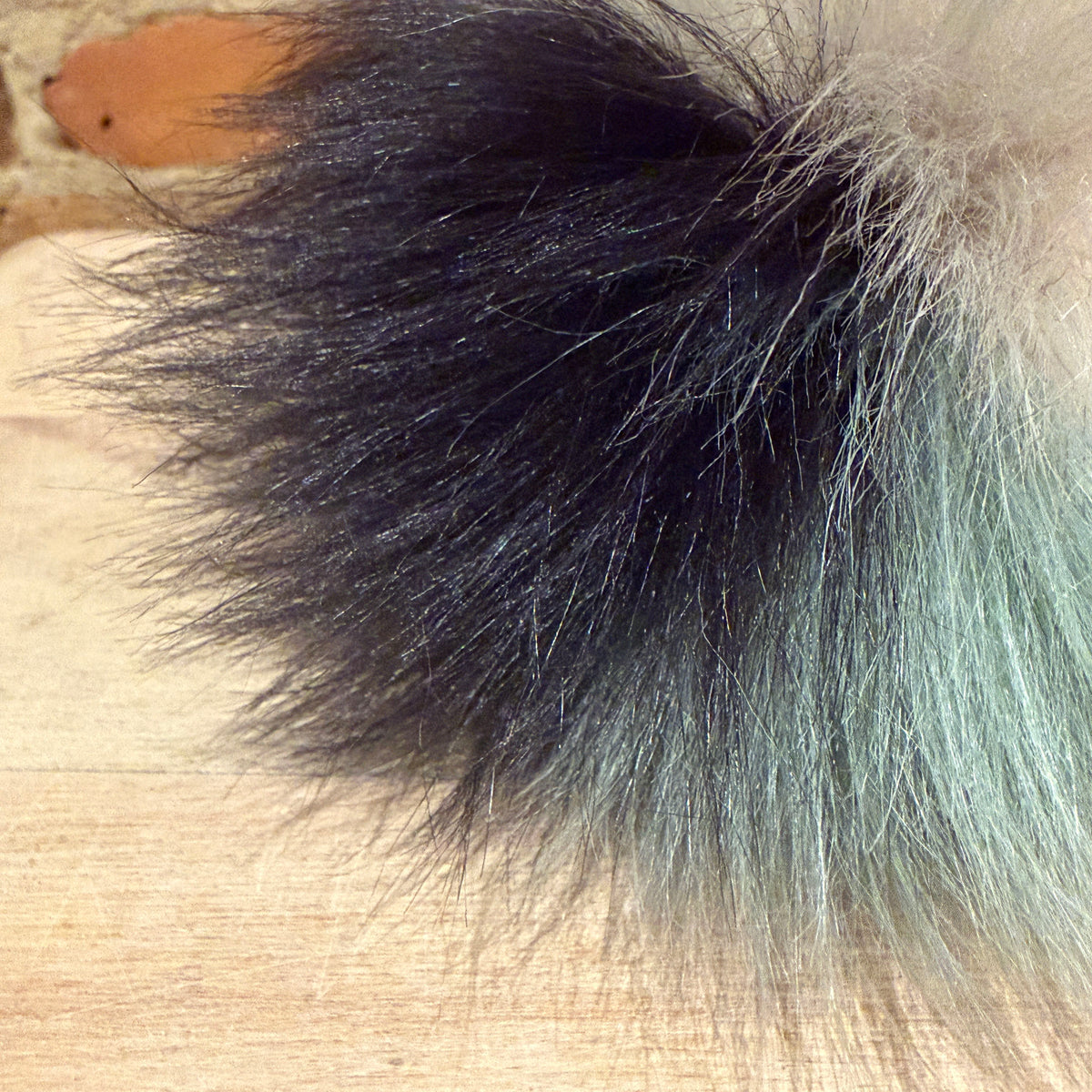 A close-up of a fluffy faux fur pom-pom accessory with a black-to-sage green gradient, resting on a light wood surface.
