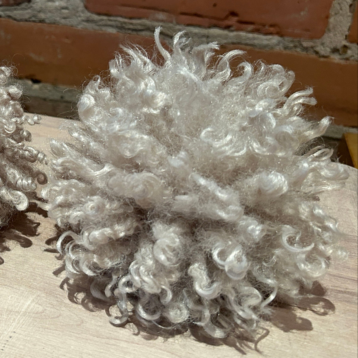 Three fluffy curly pom-poms in a light ash gray color, arranged on a light wood surface with a red brick wall background.