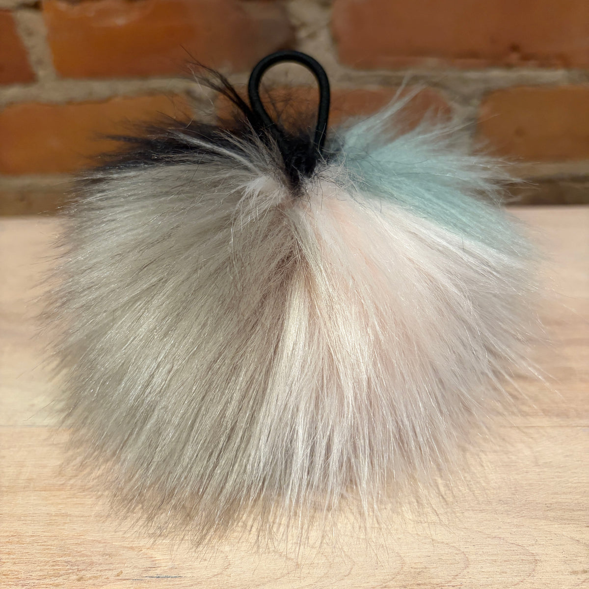A fluffy faux-fur pom pom with a black plastic loop, featuring a color-blocked design in black, dark gray, cream, pale pink, and light blue, resting on a light wood surface with a brick wall background.