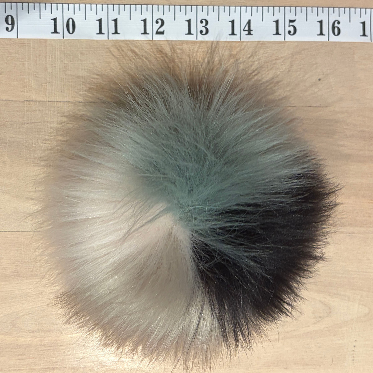 A round fluffy faux fur pom-pom with a color-blocked pattern of black, sage green, and cream, shown on a light wood surface with a measuring tape at the top for scale.