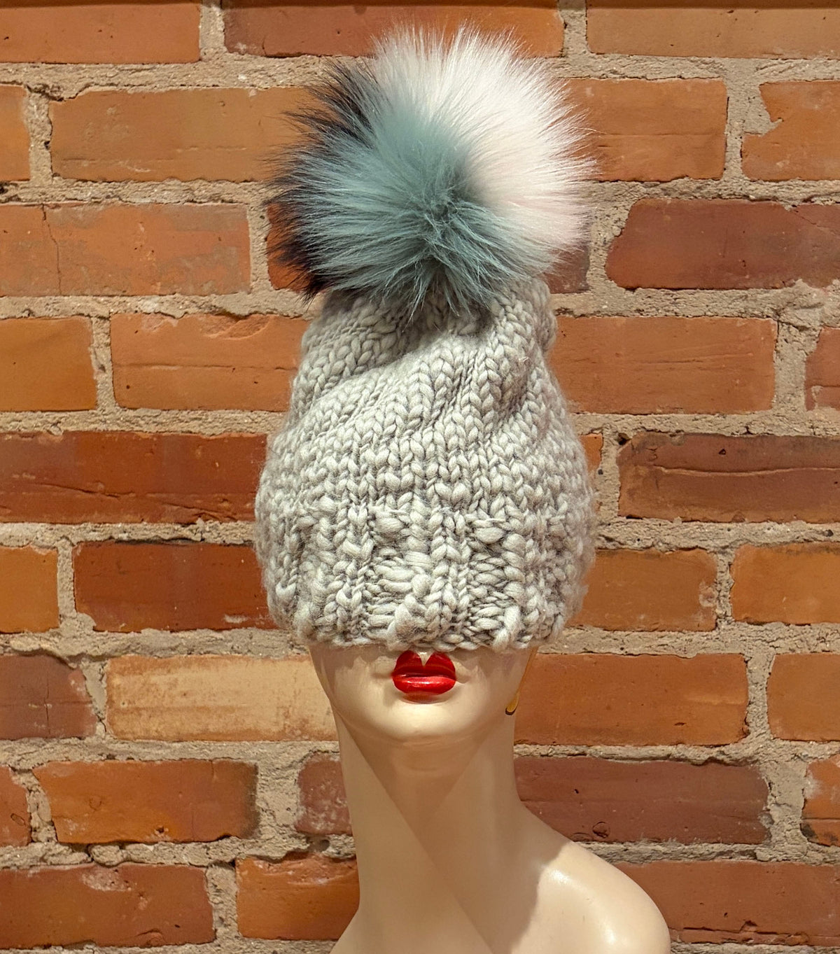 A chunky knit beanie hat in light gray with a large fluffy pom-pom in white and pale blue-gray, displayed on a mannequin head against a red-brown brick wall.