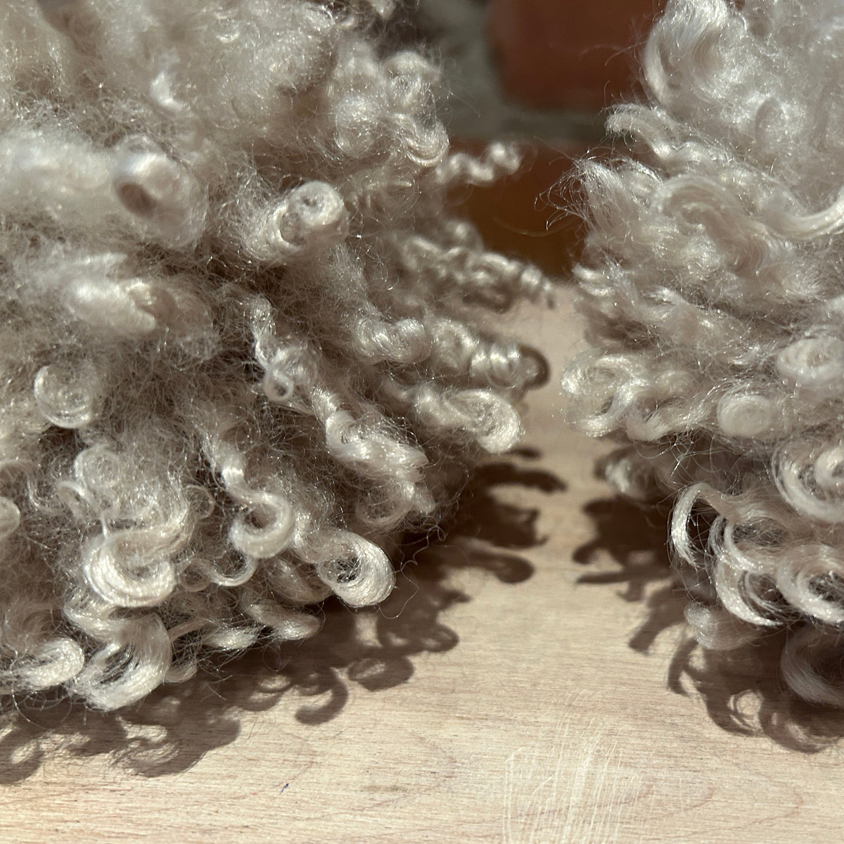 Close-up of two fluffy curly synthetic or wool-like pom-poms in a light warm gray/beige color, resting on a light wood surface.