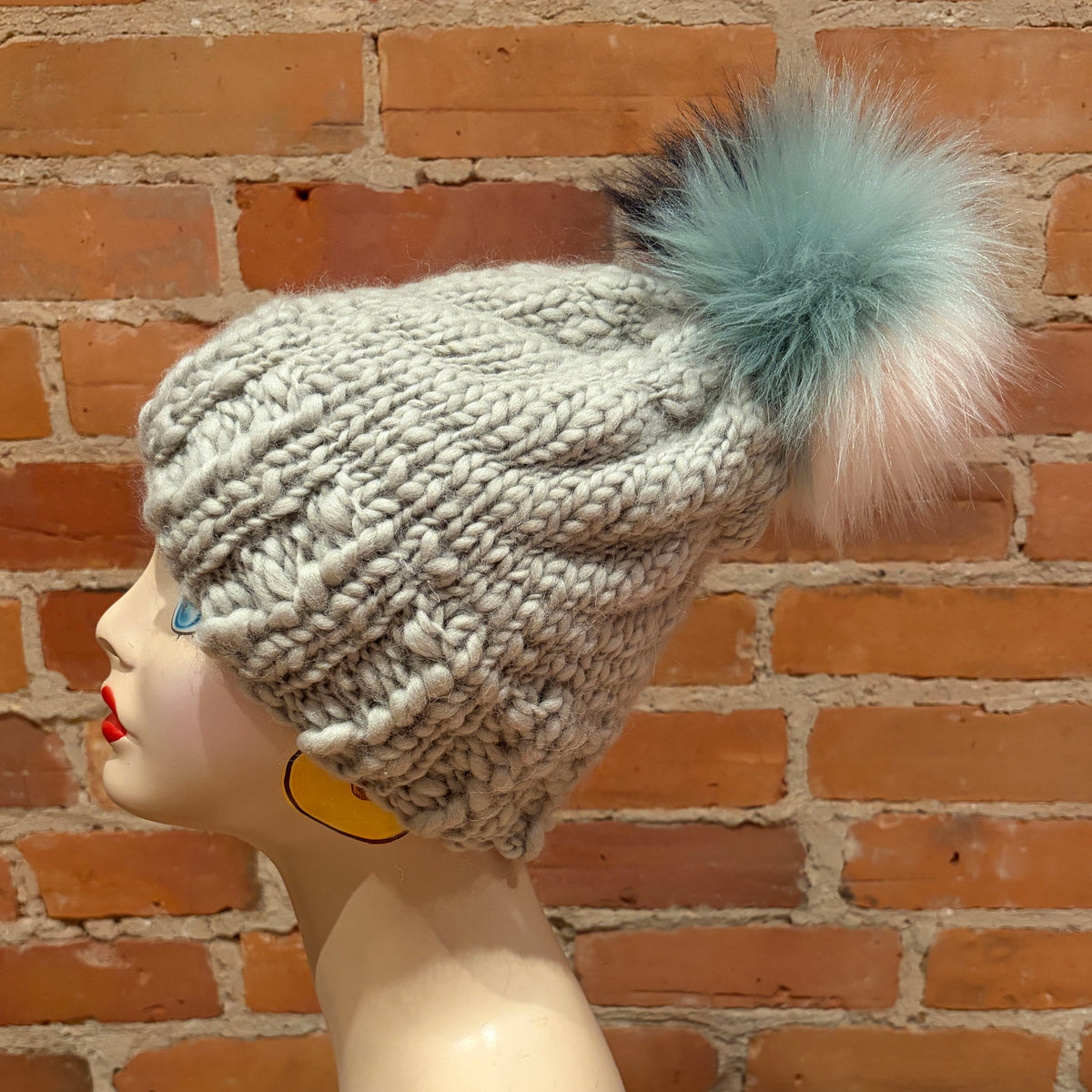 A chunky knit beanie hat in light gray with a large fluffy pom-pom in mint green, white, and pink, displayed on a mannequin head against a brick wall background.