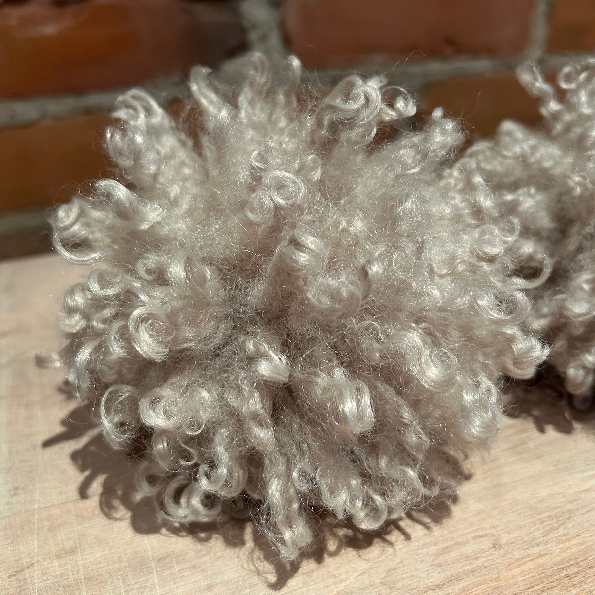 A close-up of a fluffy, curly light gray synthetic pet rug or fleece toy resting on a light wood surface with a brick wall in the background.