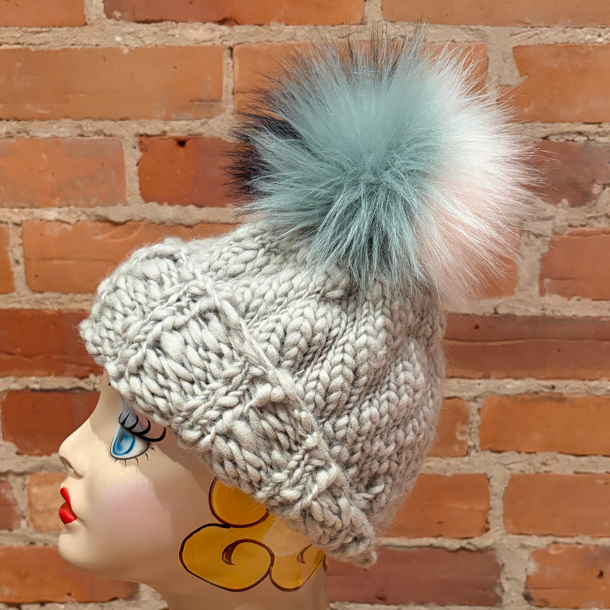 A chunky knit beanie hat in light gray with a large fluffy pom-pom in pale blue, white, and black, displayed on a mannequin head against a red brick wall background.
