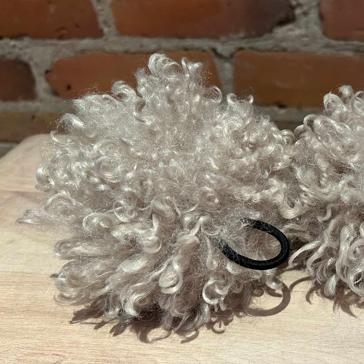 Two fluffy curly hair accessories in a light beige color, each with a black elastic loop, displayed on a wooden surface with a brick wall background.
