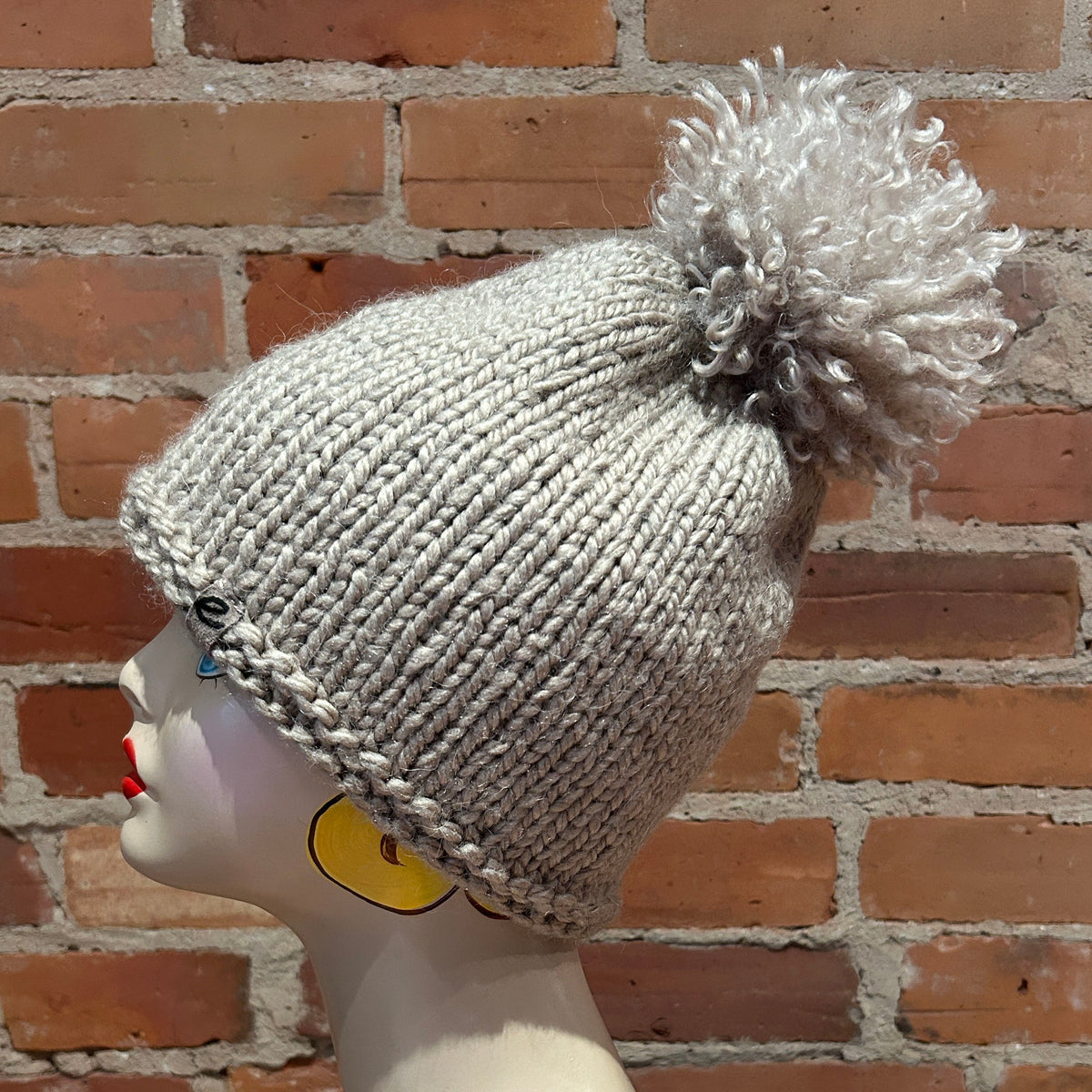 A light gray chunky knit beanie with a large fluffy pom-pom, displayed on a mannequin head against a red-brown brick wall background.