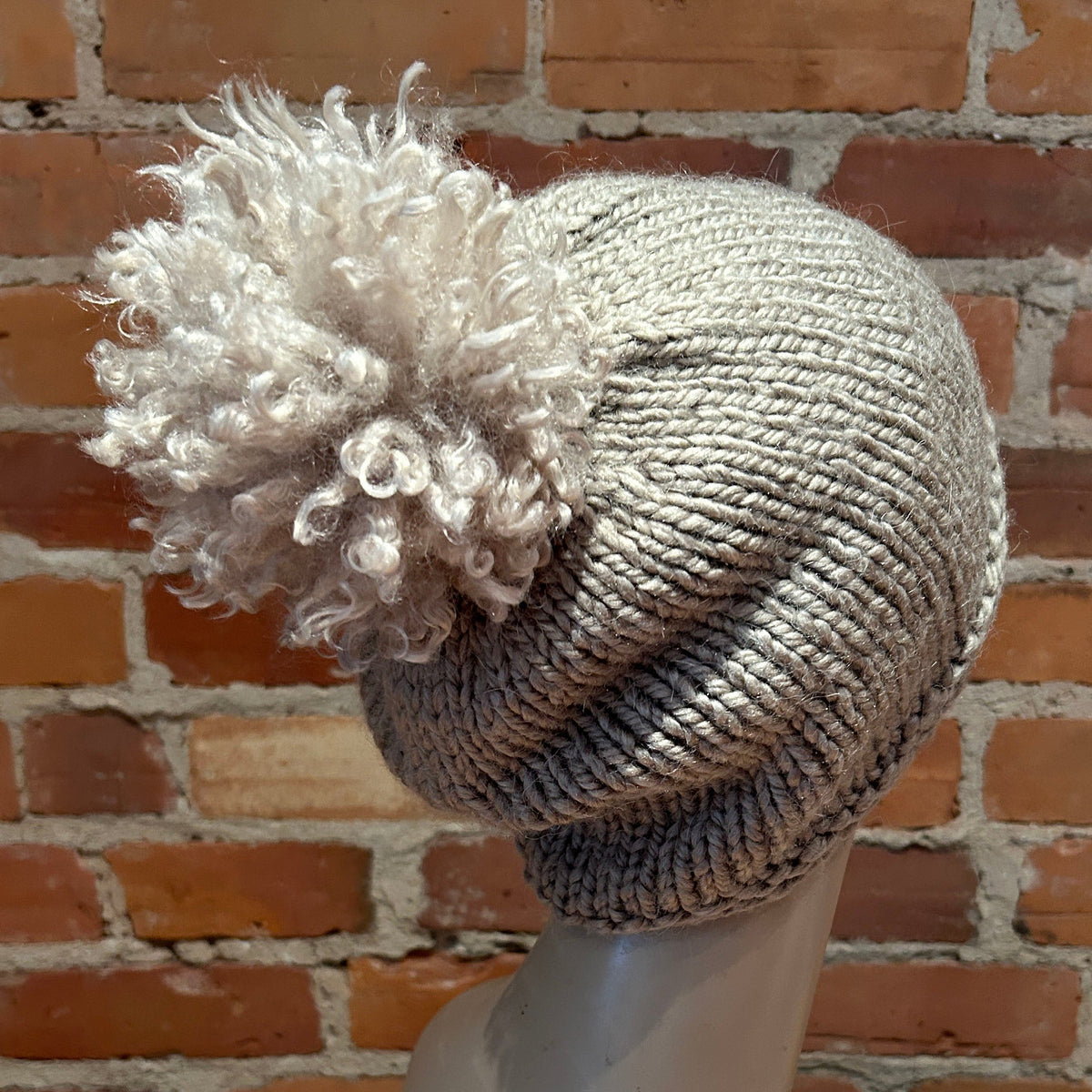 A light gray knit beanie with a large curly beige pom-pom, displayed on a mannequin head against a red brick wall background.