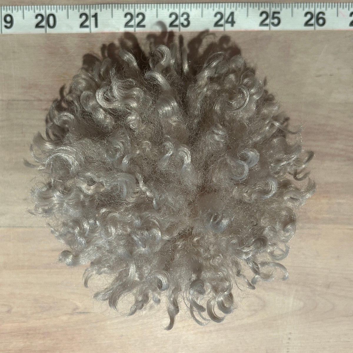 A ball of curly beige hat pom
, shown on a light wood surface with a measuring tape ruler at the top for scale.