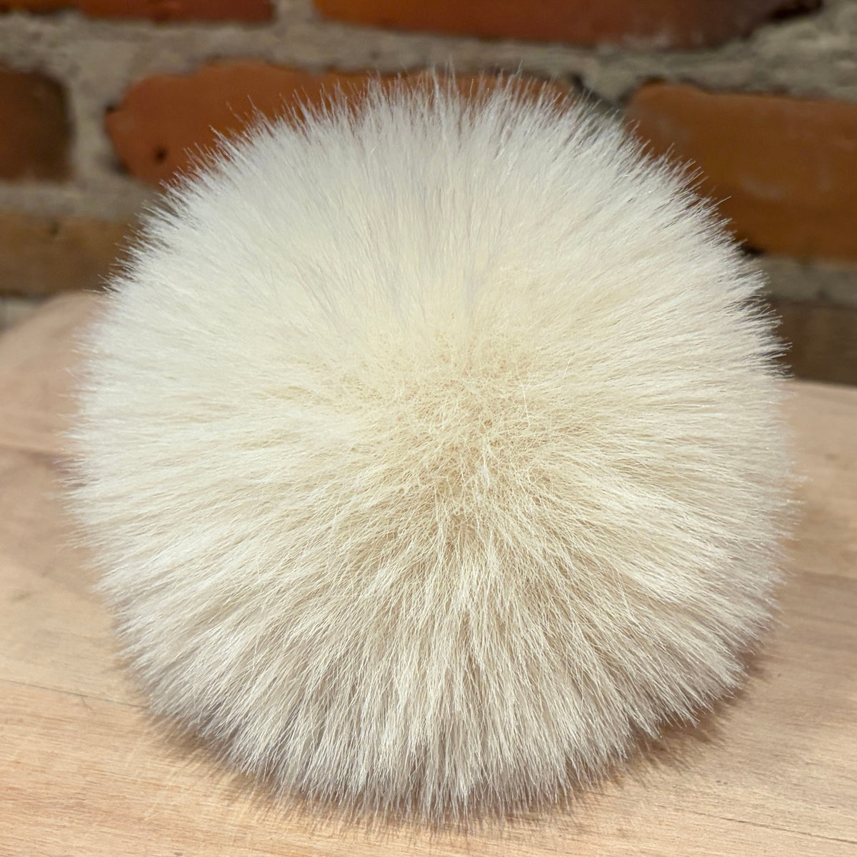 A round, fluffy pom-pom made of cream-colored faux fur, resting on a light wood surface with a blurred brick wall background.