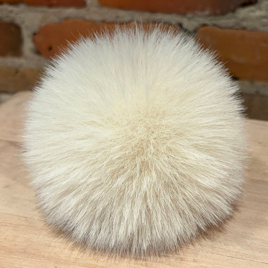 A round, fluffy pom-pom made of cream-colored faux fur, resting on a light wood surface with a blurred brick wall background.
