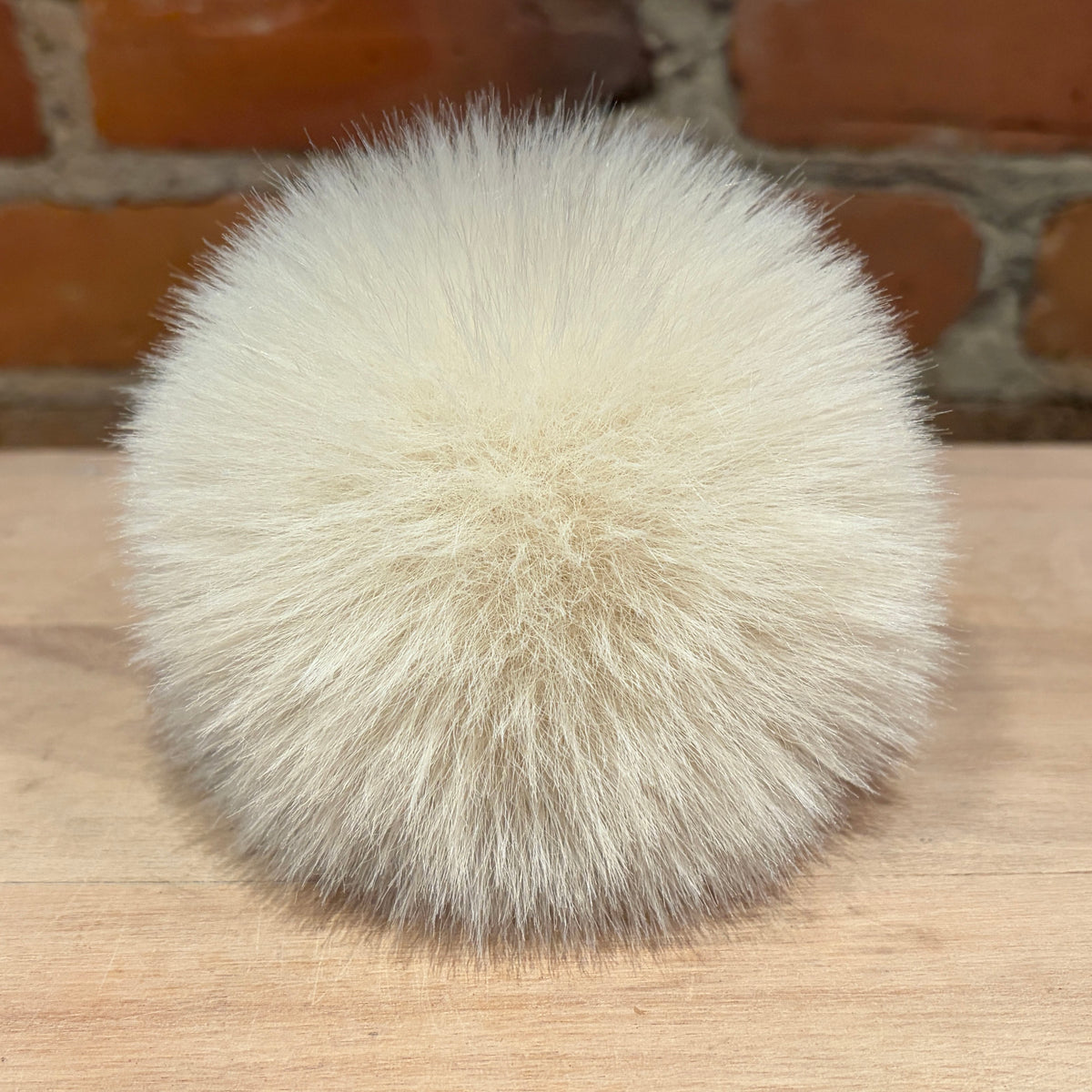 A fluffy pom-pom ball in a warm cream or off-white faux fur material, resting on a light wood surface with a brick wall background.