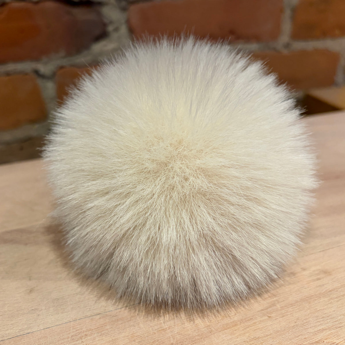 A fluffy pom-pom ball made of cream-colored faux fur or animal fur, resting on a light wood surface with a blurred brick wall in the background.