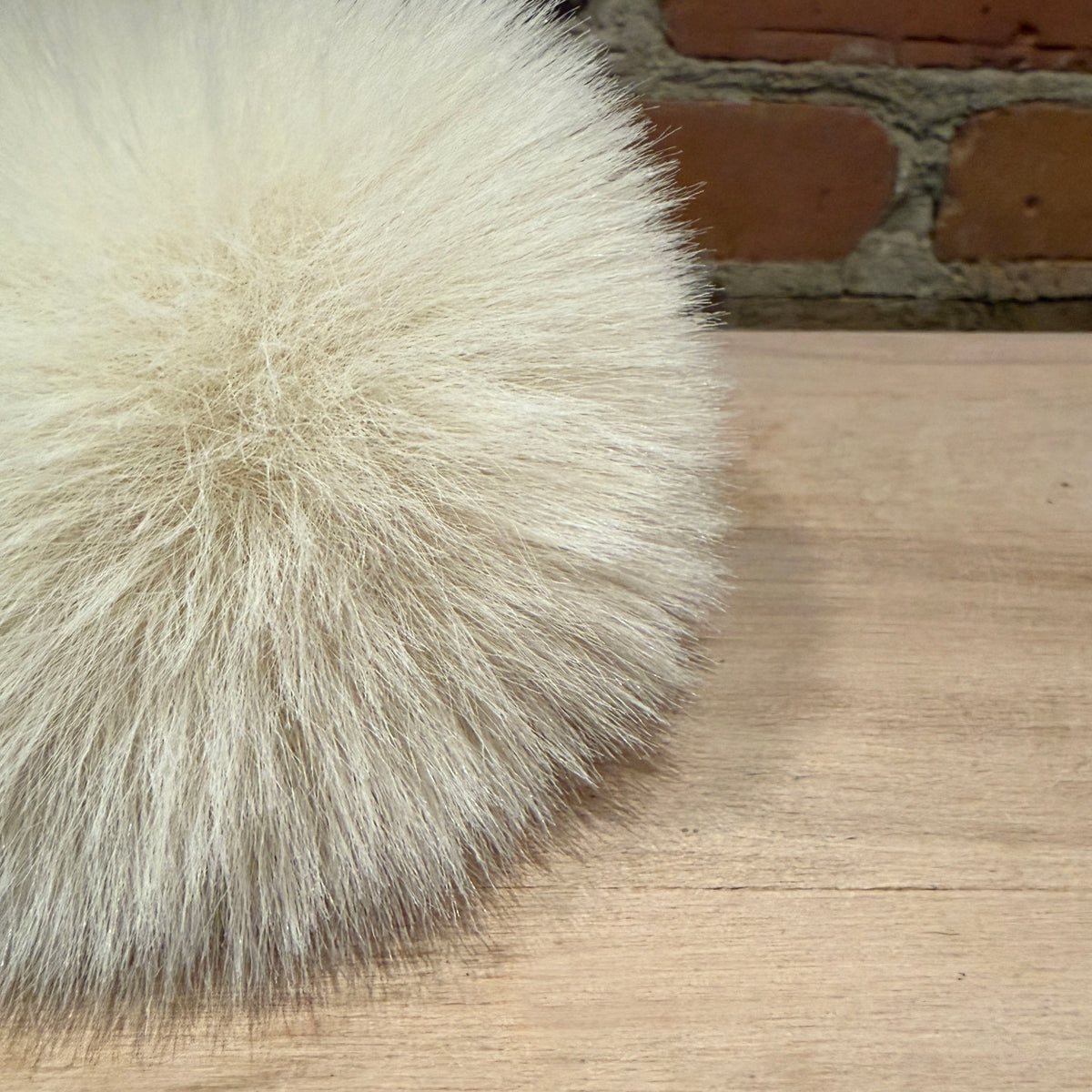 A close-up of a fluffy cream-colored faux fur pom-pom resting on a light wood tabletop with a brick wall in the background.