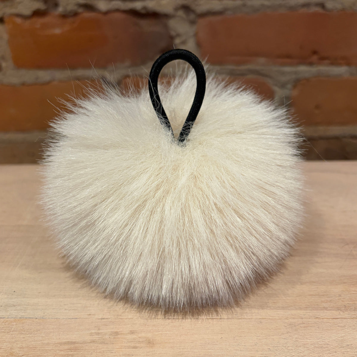 A fluffy cream faux-fur pom-pom bag charm with a black elastic loop, resting on a light wood surface with a brick wall background.