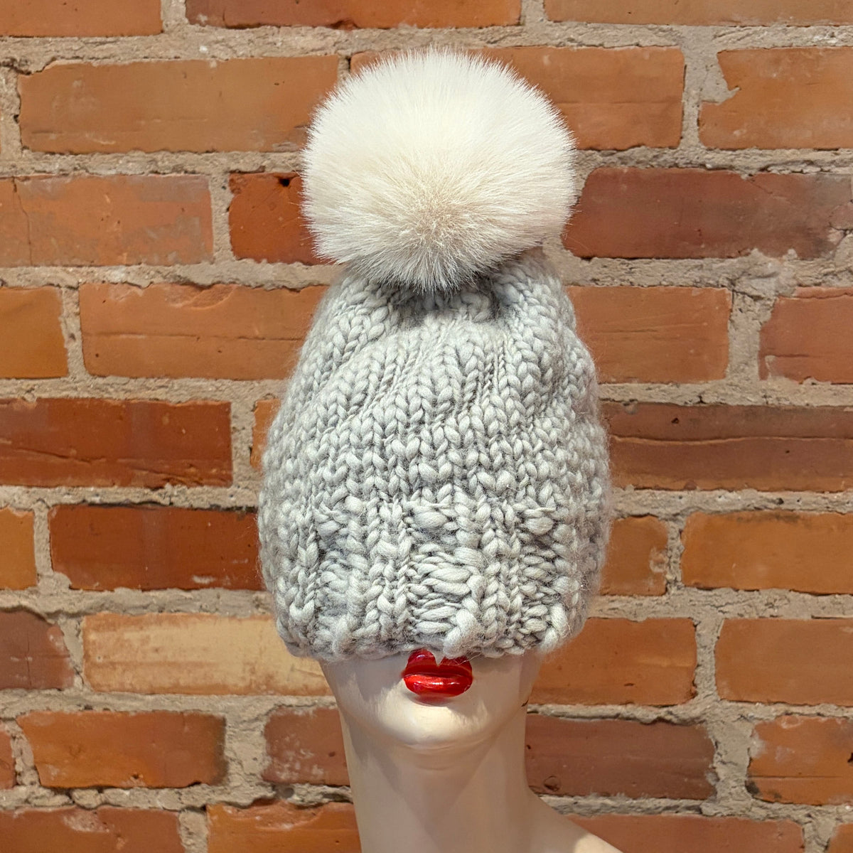 A chunky knit beanie hat in light gray with a large fluffy white pom-pom, displayed on a mannequin head against a red brick wall background.