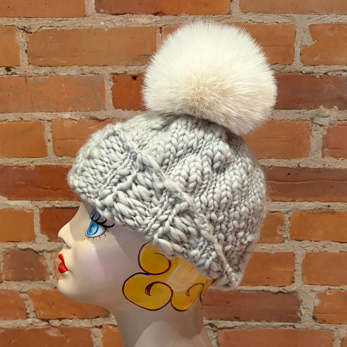A light gray chunky knit beanie with a large fluffy cream pom-pom, displayed on a decorative mannequin head against a red brick wall background.