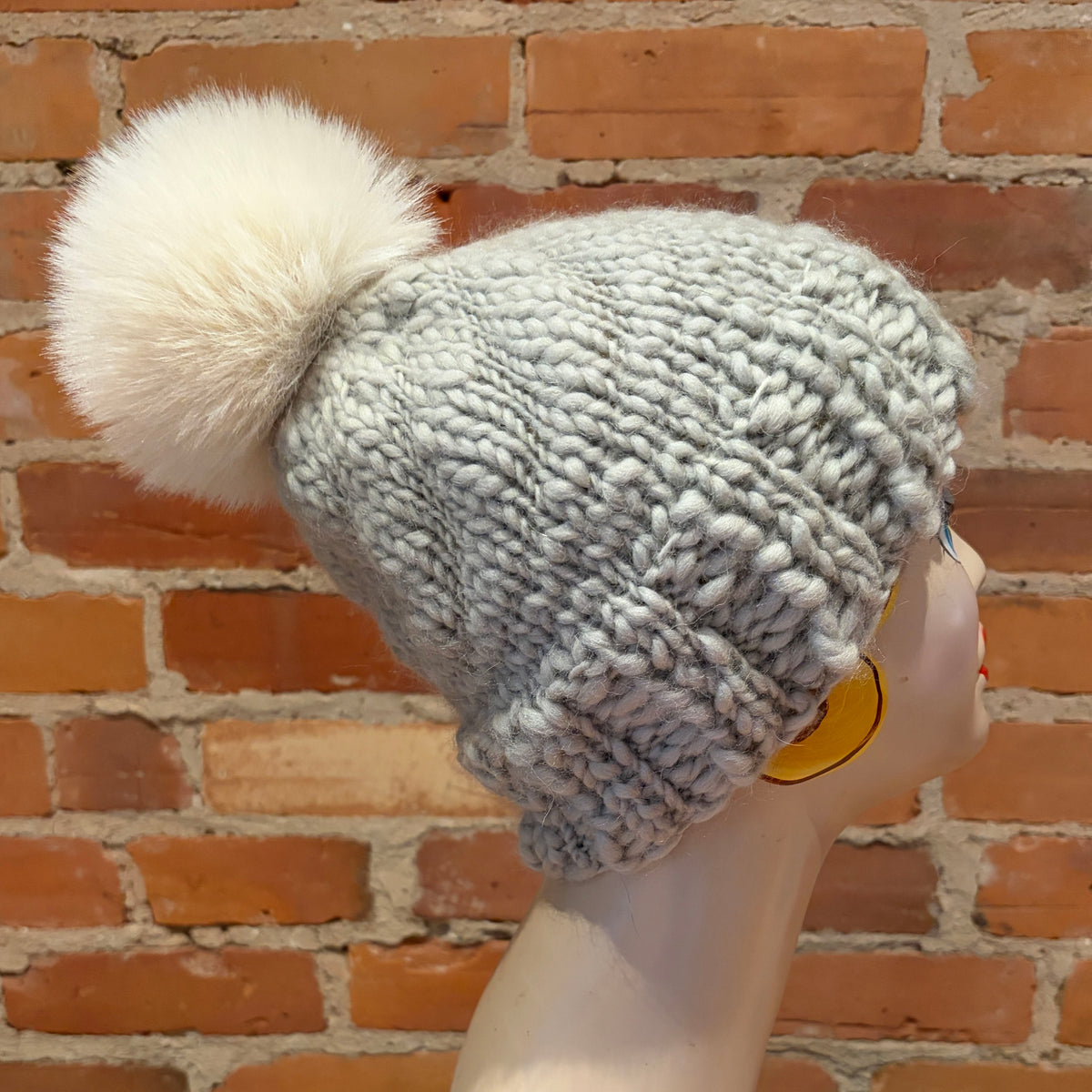 A chunky knit beanie hat in light gray with a large fluffy cream pom-pom, displayed on a mannequin head against a red brick wall background.