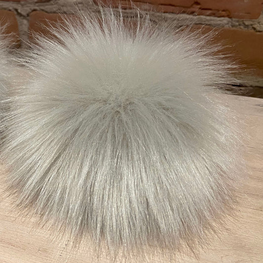 Two fluffy faux-fur pom-poms in a light silver-gray color, resting on a light wood surface with a brick wall background.