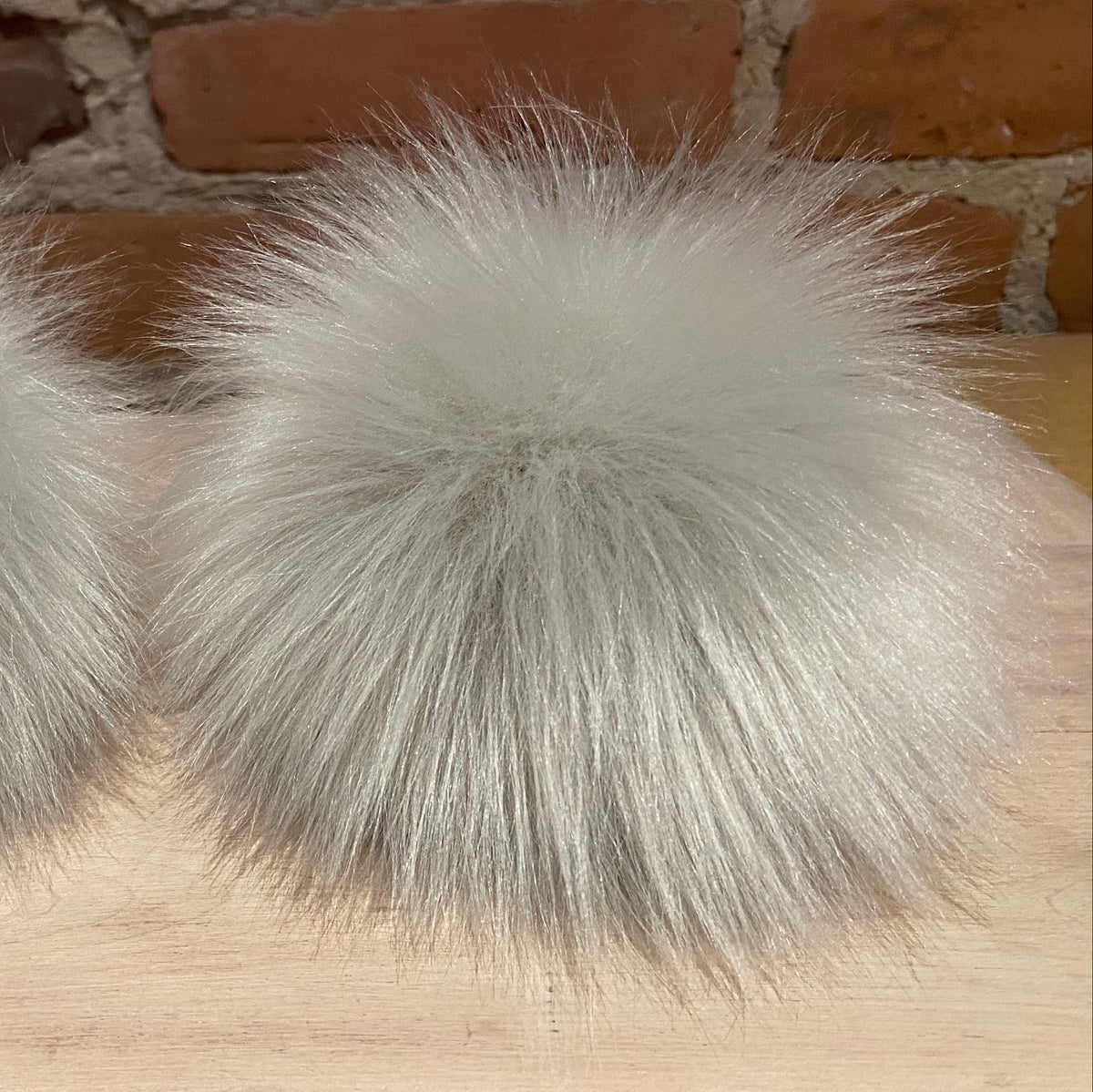 Two fluffy faux-fur pom-poms in a light silver-gray color, displayed side-by-side on a light wood surface with a brick wall background.