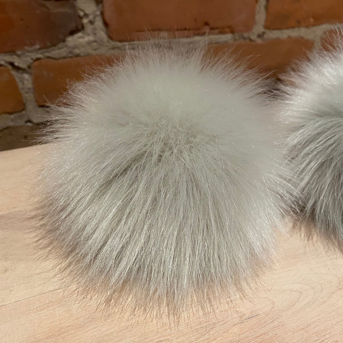 A fluffy light gray faux fur pom-pom accessory resting on a light wood surface with a brick wall background.