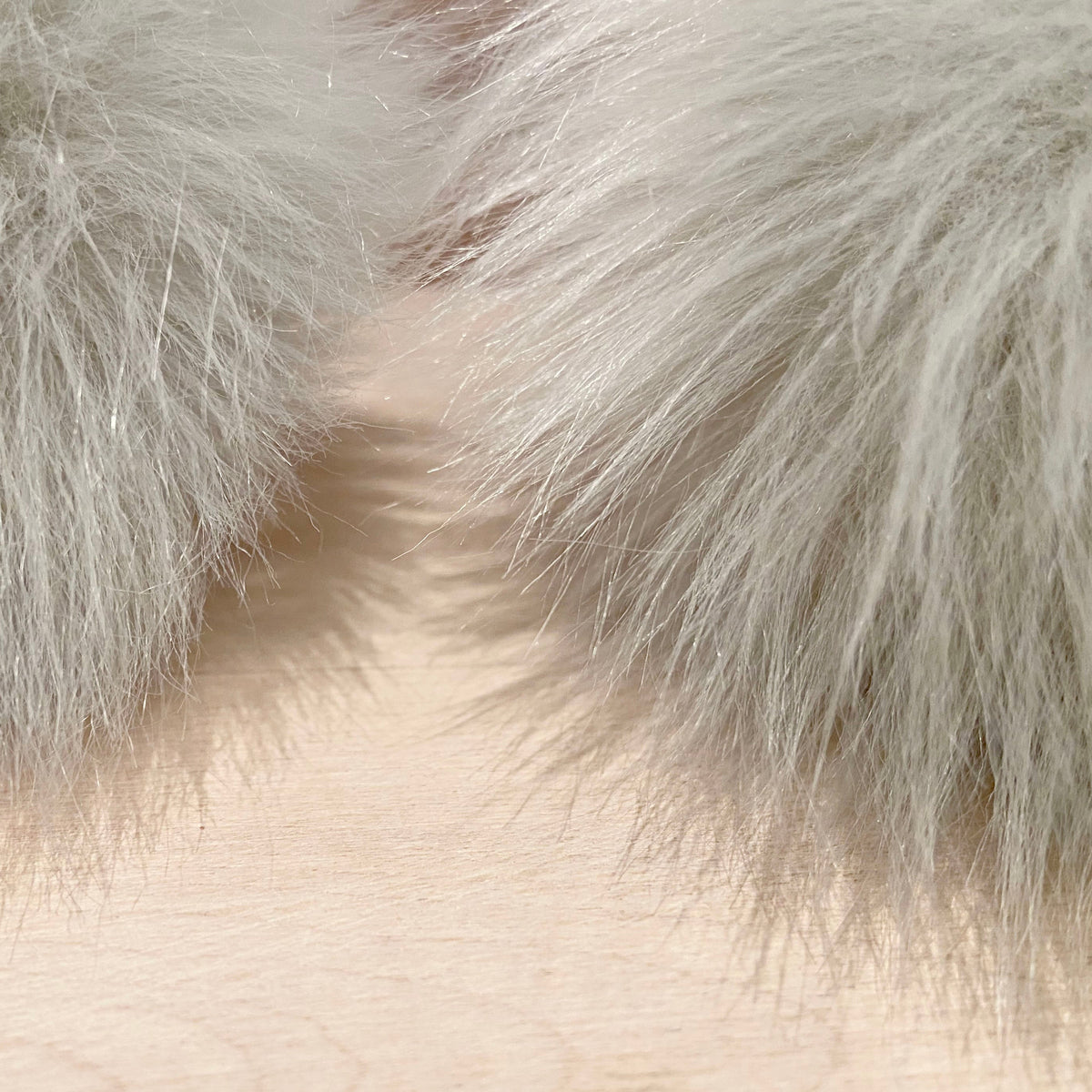 Close-up of a light gray faux fur trim or collar with soft, fluffy fibers framing a pale skin or wood-toned background.