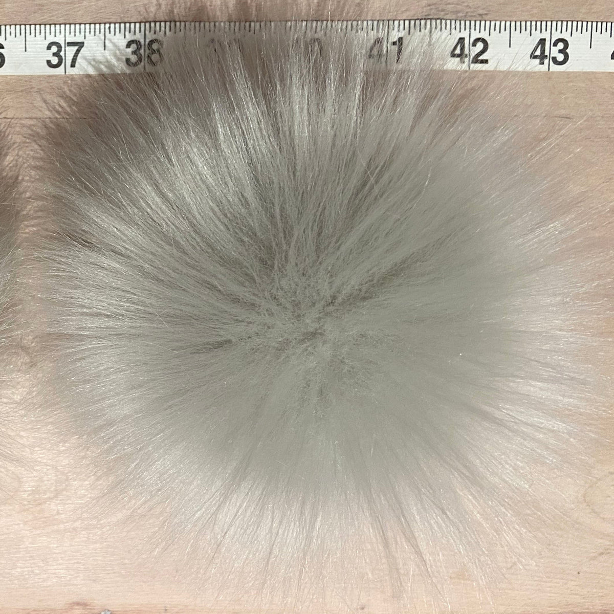 Two fluffy light gray faux fur pom-poms displayed side-by-side on a light wood surface with a measuring tape visible at the top for scale.
