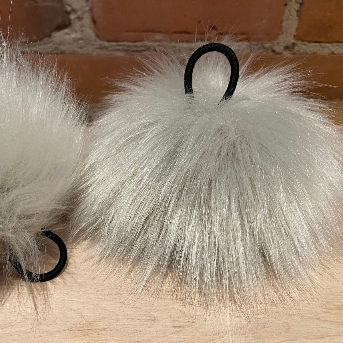 Two fluffy white faux-fur hair accessories with black rings, displayed on a light wood surface against a brick wall background.