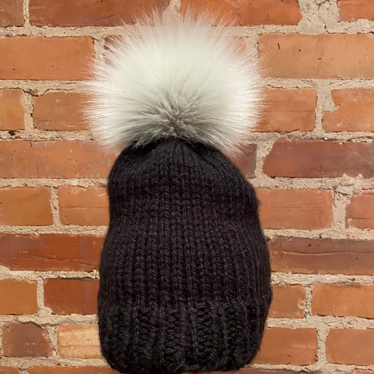 A dark charcoal gray knit beanie with a large fluffy white faux-fur pom-pom, displayed on a beige mannequin head against a red brick wall background.