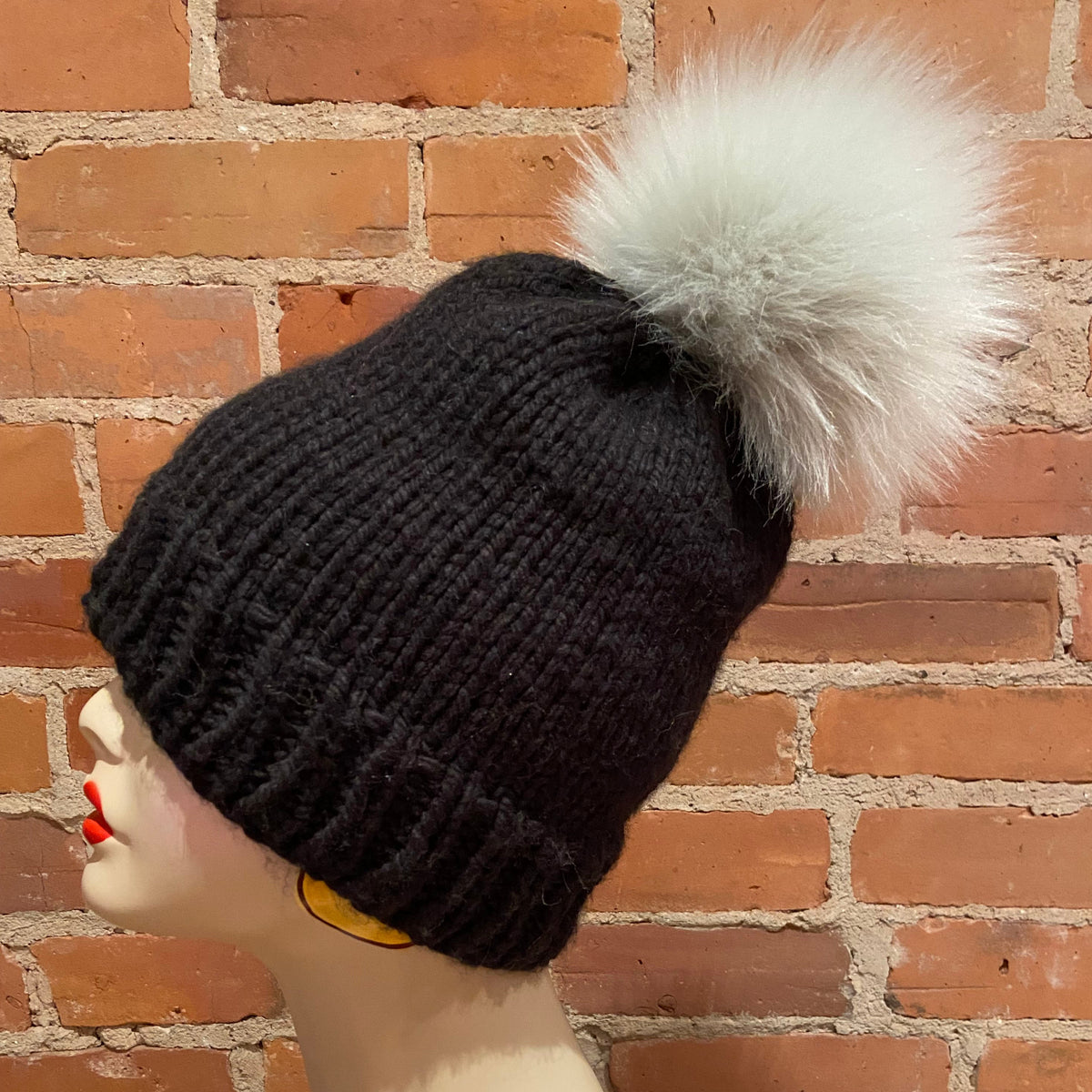 A black ribbed knit beanie with a large fluffy white faux-fur pom-pom, displayed on a mannequin head against a red brick wall background.