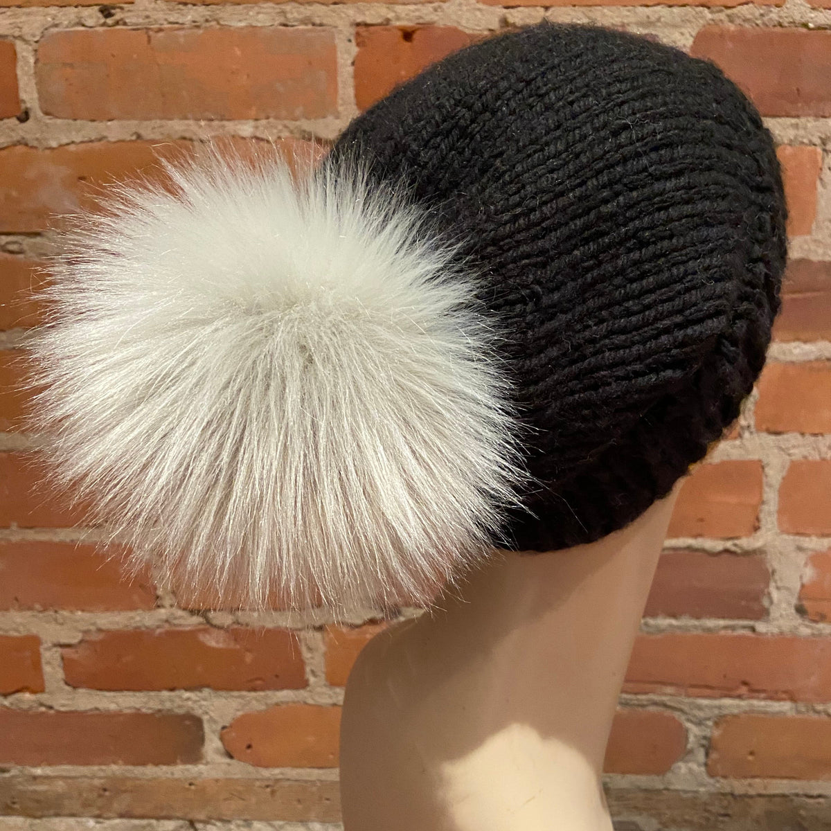 A black ribbed knit beanie hat with a large fluffy white faux-fur pom-pom, displayed on a mannequin head against a red brick wall background.