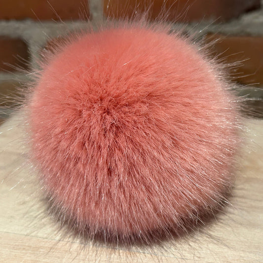 A close-up of a fluffy, dusty rose pink faux fur pom-pom accessory resting on a light wooden surface with a blurred brick wall background.