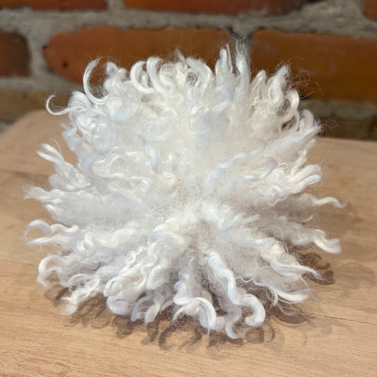 A fluffy white curly pom-pom decoration resting on a light wooden surface with a blurred brick wall in the background.