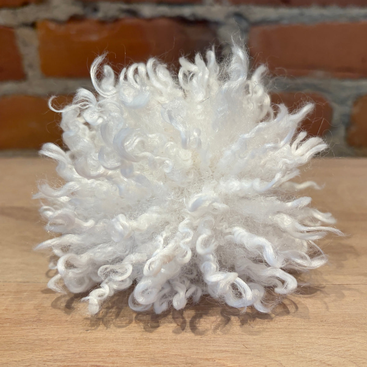 A fluffy white curly fiber ball, likely faux fur or a decorative pom-pom, resting on a light wood surface with a blurred brick wall background.