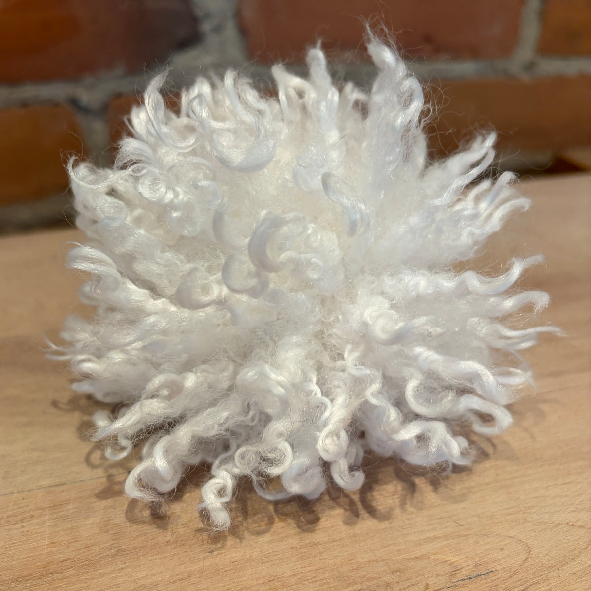 A fluffy white curly hat pom pomt, resting on a light wooden surface with a blurred brick wall in the background.