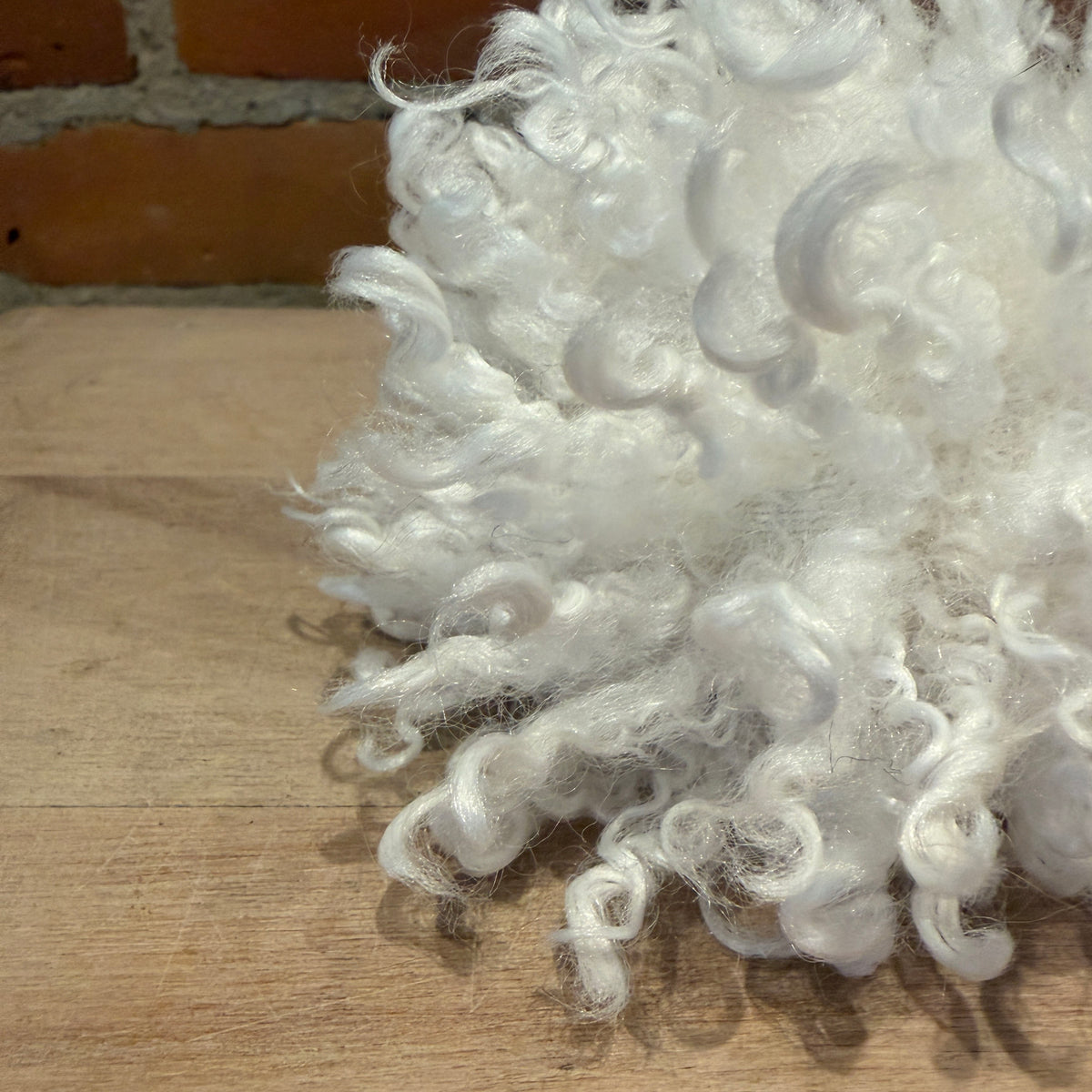 A close-up of fluffy white curly fiber, likely faux fur, piled on a light wood surface with a brick wall in the background.