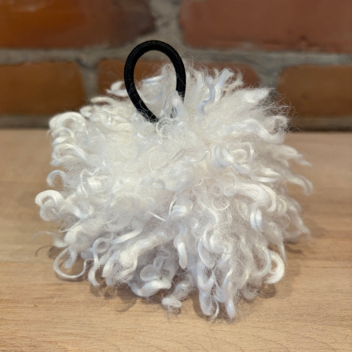 A white curly faux fur pom-pom ornament with a black elastic loop at the top, resting on a wooden surface with a brick wall background.