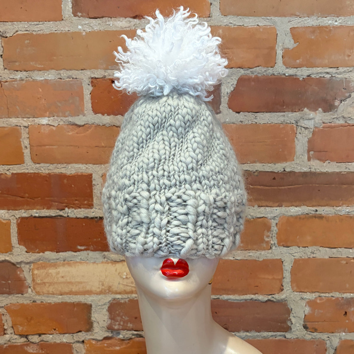 A chunky knit beanie hat in a light heather gray color with a fluffy white pom-pom on top, displayed on a mannequin head against a brick wall background.